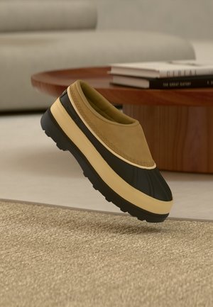 Slipper mit cognacfarbenem Obermaterial und schwarzer Gummisohle, auf beigem Teppich neben einem runden Holztisch mit gestapelten Büchern.