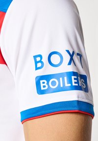 Balts sporta krekls ar ziliem un sarkaniem akcentiem, uz kura piedurknes ir zilā krāsā rakstīts "BOXT" un baltā krāsā "BOILERS". Redzama teksturēta auduma struktūra.