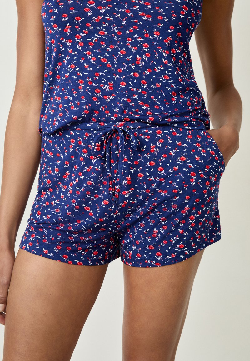 Ensemble de pyjama à motif floral avec un fond bleu marine et des fleurs rouges. Le haut a des bretelles fines, tandis que le short comprend une taille avec cordon de serrage et des poches.