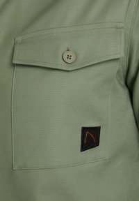 Lichtgroen shirt met een klepzak met een knoop en een zwart logopatch met oranje stiksel aan de onderkant. Glad stoftextuur.