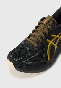 Chaussure de sport noire avec dessus en mesh, lacets vert olive, logo jaune et accents rouges. Elle présente une surface texturée et un design de semelle légère.
