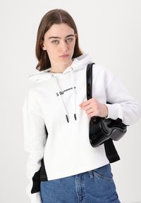 Hanorac alb cu glugă și accente negre, având un logo, șnururi și o geantă de mână netedă cu un finisaj lucios.