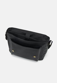 Sac croisé en cuir noir avec poche avant sécurisée par deux boutons-pression en laiton et bandoulière réglable sur fond blanc.