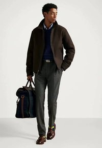 Jaunas vyras, apsirengęs tamsia striuke, tamsiai mėlynu megztiniu, pilkomis kelnėmis, rudomis loafers avalynėmis, neša didelę margą ir odinę kelionių kainą, žengia į priekį.