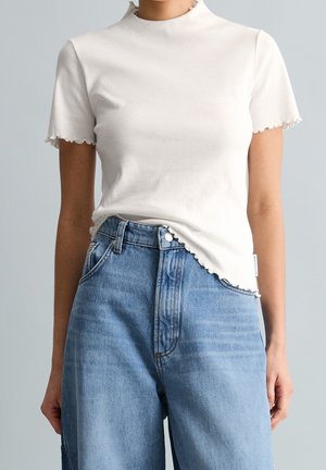 Femme portant un haut blanc côtelé à manches courtes avec des bords festonnés et un jean bleu clair taille haute sur un fond neutre.