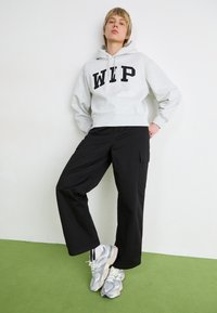 Grijze hoodie met "WIP" in vetgedrukte zwarte letters, gecombineerd met zwarte cargobroek en grijze sneakers, staand op een groene vloer tegen een witte muur.