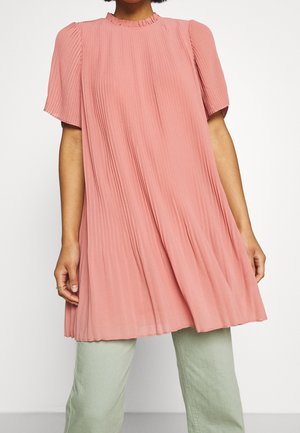 Femme portant un haut rose ample plissé à manches courtes avec un col à volants, sur un pantalon vert clair, debout devant un fond uni.