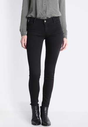 Zwarte skinny jeans met een gladde textuur, hoge taille, en subtiele zakken aan de voorkant, gecombineerd met een grijze button-up shirt en zwarte enkellaarsjes.