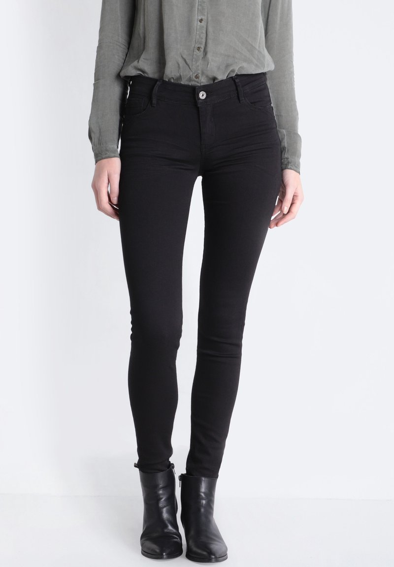 Svarta smala jeans med en slät yta, hög midja och diskreta framfickor, i kombination med en grå skjorta med knappar och svarta ankelskängor.