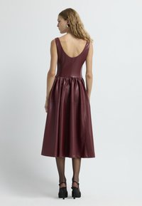 Vestido sin mangas burdeos de cuero sintético, con un escote en V profundo y falda fruncida. Combinado con tacones negros con tiras. Textura suave.