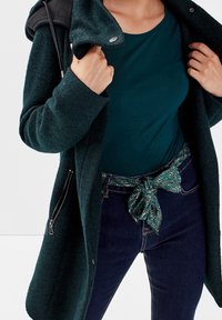 Dark green knit coat with a zippered pocket, worn over a teal top. Blue jeans featuring a teal and patterned belt tied at the waist.