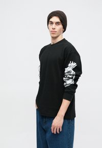 Μαύρο μακρυμάνικο μπλουζάκι με υφασμάτινη υφή, που διαθέτει λευκά γραφικά σχέδια στα μανίκια. Συνδυάζεται με μπλε τζιν παντελόνι. Φορεμένο καφέ σκουφάκι.