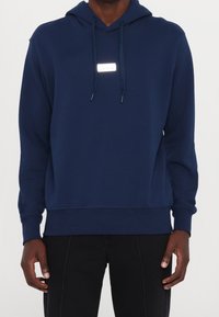 Marineblauer Kapuzenpullover aus weichem Stoff, mit einem Logo-Patch vorne, verstellbarer Kapuze mit Kordelzug sowie gerippten Bündchen an Ärmeln und Saum.