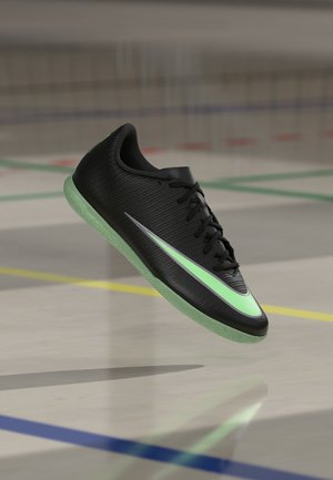 Chaussure de sport d’intérieur noire avec semelle verte lumineuse et grand swoosh, flottant au-dessus d’un sol de gym marqué de lignes colorées.