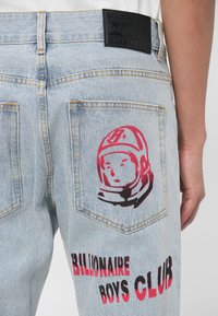 Jean en denim bleu clair avec un patch en cuir noir, présentant un graphique d'astronaute rouge sur la poche arrière et le texte "BILLIONAIRE BOYS CLUB".