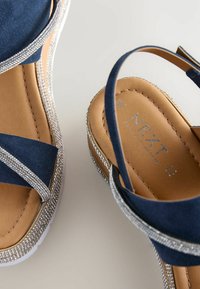 Sandales en daim bleu marine avec un accent argenté scintillant, une semelle plate et un design à lanières croisées. Comprend une semelle intérieure rembourrée marron avec un logo.