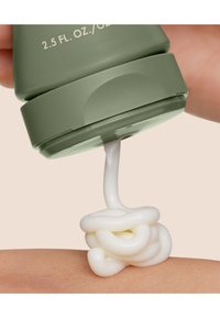 Grønn klemmetube som doserer kremet hvit lotion på huden. Teksturen virker glatt og tykk, med en subtil glans. Tuben viser produktets størrelse.