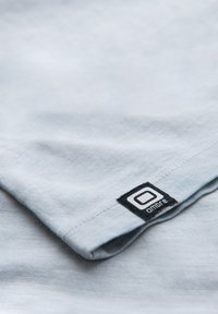 Světle modré bavlněné tričko s texturovaným povrchem, na kterém se nachází černá nášivka v dolním rohu. Design je jednoduchý s kulatým výstřihem.