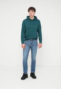 Φούτερ με κουκούλα σε πετρόλ χρώμα με κορδόνια και μπροστινή τσέπη· τυπωμένο λογότυπο στο στήθος. Τζιν παντελόνι σε ανοιχτό μπλε και μαύρα αθλητικά παπούτσια συμπληρώνουν το ντύσιμο.