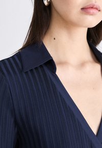 Navy blue top with vertical pleats, a pointed collar, and a deep V-neck design. Fabric appears smooth with a structured fit. Silver earring visible.