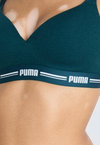 Teal sports-bh med V-halsdesign. Har en bred, elastisk bånd med hvit "PUMA"-logo og kontrasterende striper. Glatt stofftekstur.