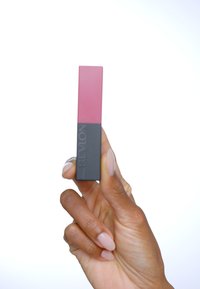 Produkt do ust Revlon w prostokątnej opakowaniu, z różową górną częścią i czarną dolną częścią, trzymany w dłoni na białym tle.
