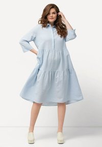 Vestido azul claro, en capas, hecho de una tela suave y texturizada. Cuenta con un frente abotonado, cuello y mangas de tres cuartos, con una silueta fluida.