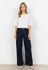 Vit kortärmad topp med broderade detaljer och mörkblå vida jeans. Jeanserna har två fickor och beige sömmar längs kanterna.
