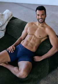 Men's navy boxer briefs with a red waistband, sitting on a green textured couch. The model has a toned physique and is relaxed.