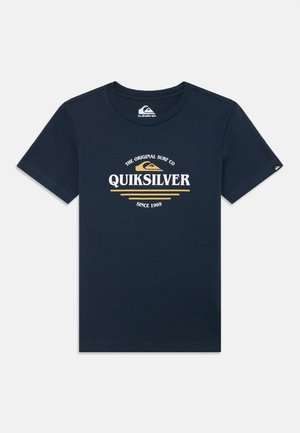 Marineblå bomull t-skjorte med korte ermer. På forsiden vises "Quiksilver"-logoen i hvitt og gull, med buet tekst og dekorative linjer.