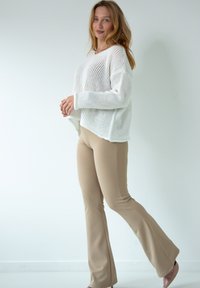 Biały sweter z dzianiny o luźnym kroju i prążkowanej fakturze, zestawiony z beżowymi spodniami dzwonami. Modelka stoi w profilu, prezentując outfit.
