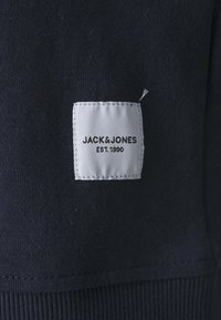 Marine stof met een wit vierkant label waarop "JACK & JONES EST. 1990" in zwarte tekst staat, met gestikte randen en een gladde textuur.