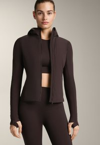 Brun zip-up jacka med huva, figursydd design, långa ärmar med tumhål, parat med matchande högmidjade leggings. Slät textur.