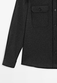 Camicia in lana grigia scura con bottoni, maniche lunghe e tasca sul petto con patta e bottone su sfondo bianco.