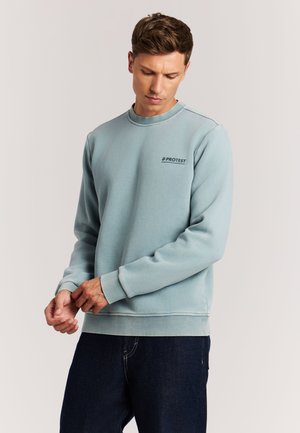 Man in lichtblauwe trui met 'PROTEST' logo die zijn mouw aanpast, draagt een donkere spijkerbroek, staande tegen een effen lichte achtergrond.