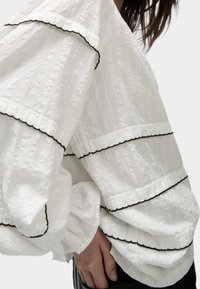 Blusa blanca texturizada con volantes en capas con ribete negro, mangas abullonadas y puños ajustados, destacando detalles de diseño únicos.