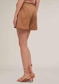 Ruskean mokkanahkashortsit vyöllä, joissa on muotoiltu malli ja käänteet lahkeissa. Yhdistetty tanväreisiin remmisandaaleihin neutraalilla taustalla.