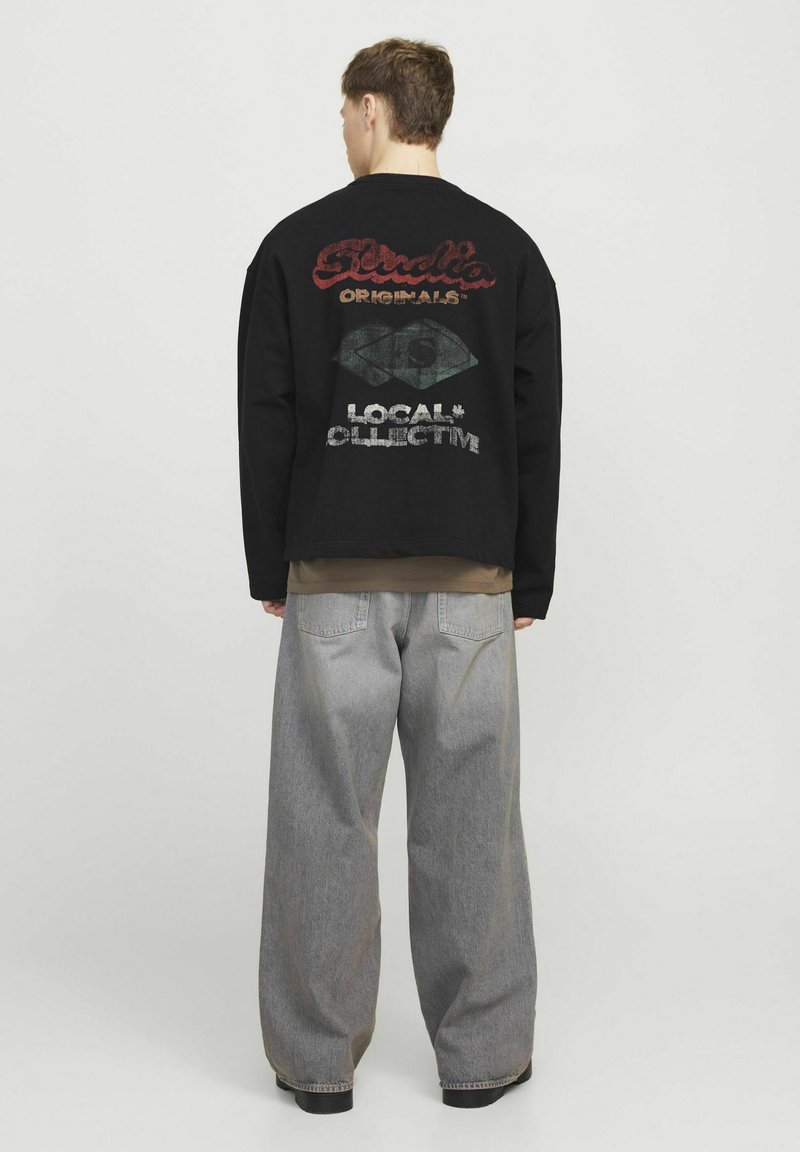 Felpa nera oversize con grafiche rosse e verdi sul retro, abbinata a pantaloni in denim grigi larghi. Texture liscia.