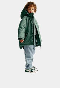 Zelená vycpaná parka s kapucí, s ferměn�ím zipem a tlačítkovým zapínáním, spárovaná se světle modrými džínami a teniskami s zelenými akcenty.