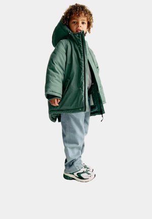 Grüne gepolsterte Parka mit Kapuze, ausgestattet mit einem Reißverschluss und Druckknopfverschluss, kombiniert mit hellblauen Jeans und Turnschuhen mit grünen Akzenten.