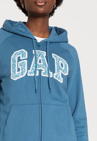 Felpa blu con zip, con la scritta "GAP" in stampa floreale bianca, cordino e texture morbida. Tasche a marsupio sul davanti.