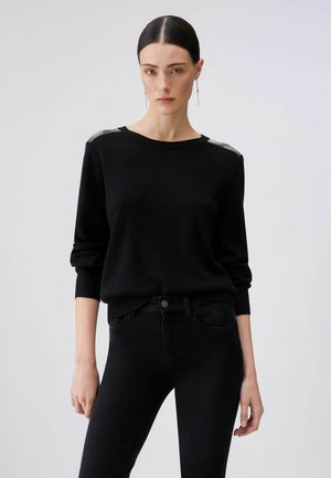Donna con capelli lisci pettinati all'indietro, che indossa un maglione nero con dettagli in strass sulle spalle e pantaloni neri, in piedi contro uno sfondo chiaro e semplice.