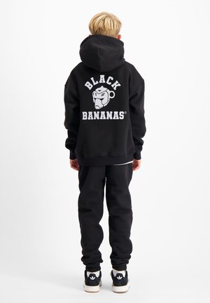Kind met blond haar, gekleed in een zwarte hoodie en broek, met de rug naar de kijker; op de hoodie staat witte tekst "Black Bananas" en een apenlogo op de rug.