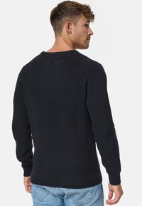 Pull en tricot noir avec texture côtelée, cols ronds et manches longues. Matière douce, design ajusté. Porté avec un jean en denim bleu.