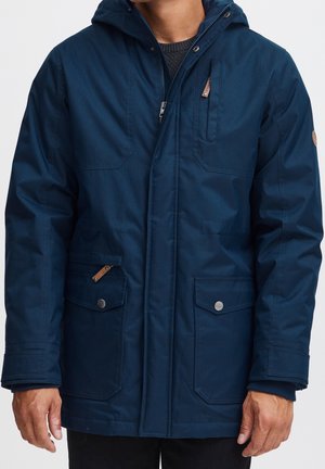 Mężczyzna w granatowej zimowej kurtce z kapturem, zapinanej z przodu na zamek, z dwiema kieszeniami na zatrzaski oraz kieszenią na piersi zapinaną na zamek.