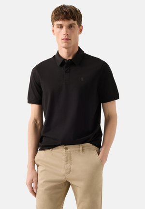 Jonge man die een effen zwart poloshirt met korte mouwen en beige broek draagt, staand met één hand in de zak tegen een effen achtergrond.