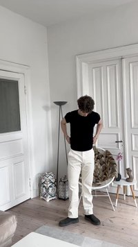 Sort kortærmet t-shirt, hvide bukser og sort fodtøj. Moderne interiør med trægulv, dekorative potter og en gulvlampe.