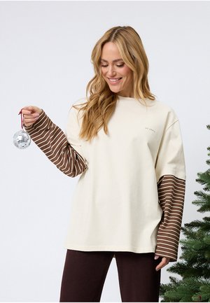 Krémově zbarvená mikina s dlouhými hnědými pruhovanými rukávy, s kulatým výstřihem a malým logem na hrudi. Drží ozdobný předmět.