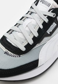 Tenisky Puma s šedým textilním svrškem, černými semišovými akcenty, bílou koženou boční panelem a plochými tkaničkami. Obsahují viditelné švy a logo.