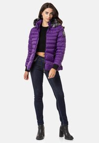 Lila Puff-Jacke mit einem gefütterten Kapuzenkragen, Reißverschluss vorne, gerippten Bündchen und einem Patch-Logo. Kombiniert mit dunklen Jeans und schwarzen Stiefeletten mit Absatz.